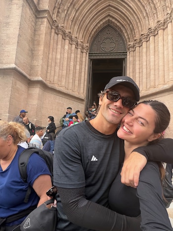 Una mujer y un hombre sonríen abrazados frente al portal de una basílica de piedra con personas de fondo