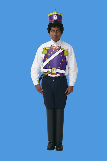 Un joven de pie sobre fondo azul lleva camisa blanca, pantalón oscuro y una pechera con sombrero de papel
