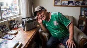 Hombre de mediana edad, con camiseta verde de México y gorra, escucha una radio vintage con la mano en la oreja en una habitación con una ventana y fotos familiares.