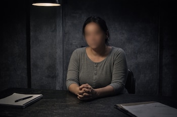 Mujer con rostro difuminado sentada en una mesa oscura y manos entrelazadas, bajo una lámpara cenital en una sala con paredes texturizadas; hay un cuaderno y papeles.