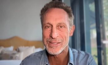 Hombre de mediana edad con cabello castaño y canas, barba incipiente, camisa de jean, mirando al frente y sonriendo. Fondo interior con cabecero y ventana