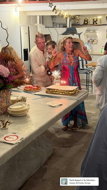 Un hombre sostiene a un niño junto a una mujer con vestido colorido que celebra frente a una tarta iluminada por bengalas sobre una mesa