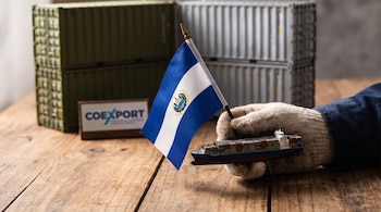 Mano con guante sostiene una pequeña bandera de El Salvador y una maqueta de buque de carga sobre una mesa de madera con contenedores.