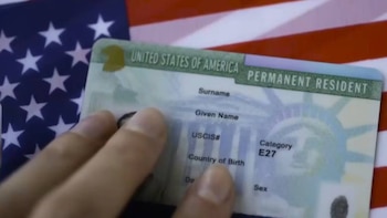 Una mano sostiene una Tarjeta Verde frente a la bandera estadounidense. La tarjeta muestra "UNITED STATES OF AMERICA PERMANENT RESIDENT" y campos de texto