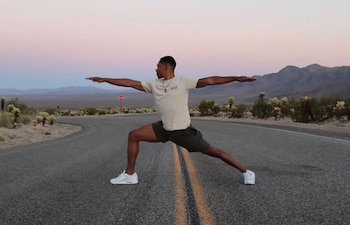 Hombre practicando yoga en un tapete azul junto a un río verde. Viste camiseta beige y shorts azules. Al fondo, edificios coloridos y montañas