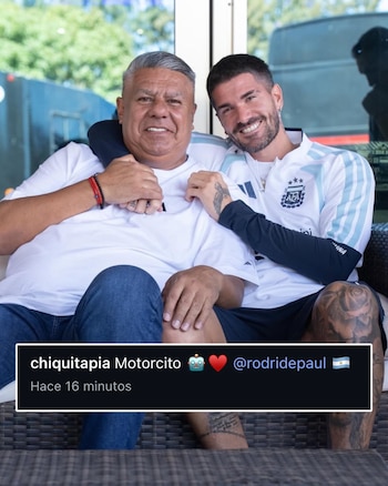 Claudio Tapia, de cabello gris y camiseta blanca, abraza a Rodrigo De Paul, de barba y chaqueta celeste y blanca, ambos sonriendo sentados