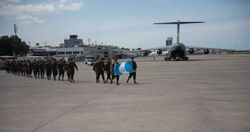 Militares uniformados caminan en una pista de aeropuerto. Algunos portan la bandera de Guatemala. Un gran avión de carga y edificios de aeropuerto visibles