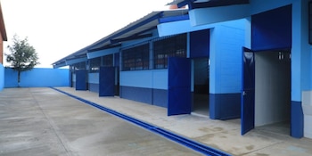 Vista exterior de un edificio escolar moderno con paredes azules y oscuras, un techo metálico, ventanas con rejas y puertas abiertas hacia aulas. Un patio de concreto con un desagüe azul y un árbol al fondo