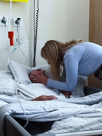 Evelyn Von Brocke se inclina sobre un hombre en una cama de hospital, sujetando su rostro. Se observa equipo médico y sábanas blancas en el entorno hospitalario