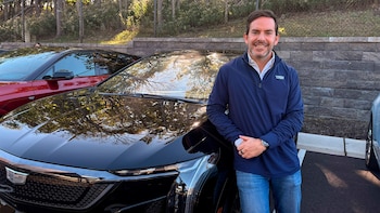 Un hombre sonríe apoyado junto a un automóvil negro de la marca Cadillac en un estacionamiento al aire libre