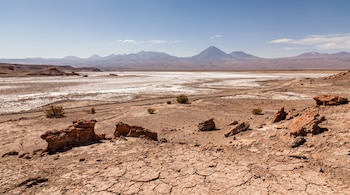 Vista panorámica del desierto de Atacama con tierra agrietada, rocas rojizas, salares blancos, un camino de tierra y montañas volcánicas bajo un cielo azul claro.