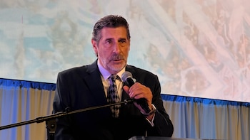 Hombre de mediana edad con barba y traje oscuro, sujetando un micrófono y hablando. Fondo con pantalla LED y cortinas blancas y azules