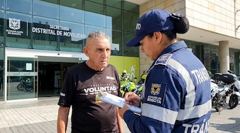 Agente de tránsito de Bogotá en uniforme azul oscuro y gorra escribe en un portapapeles a un hombre, con la Secretaría de Movilidad de fondo.
