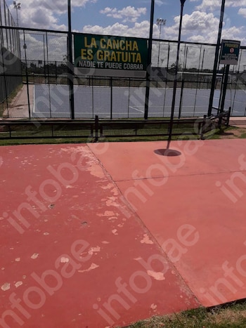 Cancha deportiva cercada. Un cartel verde indica que el acceso es gratuito. Otro cartel prohíbe bebidas alcohólicas. Primer plano de suelo rojo con desgaste
