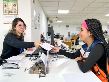 Dos mujeres intercambian documentos en un escritorio de oficina. Una usa gafas y la otra una diadema rosa y chaleco naranja. Se ven monitores y material de oficina