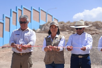 Tres personas cortan con tijeras una cinta roja y blanca frente a un muro escalonado y un terreno de tierra bajo la luz del sol