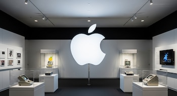 Un logotipo blanco de Apple iluminado en una sala de exposición, rodeado por vitrinas con computadoras y dispositivos antiguos de la marca.