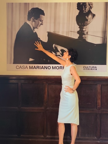 Cecilia Muilone, vestida de blanco, sonríe y extiende su mano hacia un gran póster en blanco y negro de Mariano Mores tocando el piano