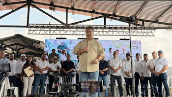 Alfonso Portillo, vestido con camisa beige, habla en un micrófono frente a un grupo de personas y una pantalla grande con imágenes