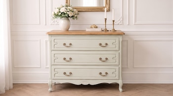 Cómoda de tres cajones con tapa de madera. Sobre ella, jarrón de rosas blancas, dos candelabros con velas, una caja. Espejo con marco dorado. Pared blanca y suelo de parquet.