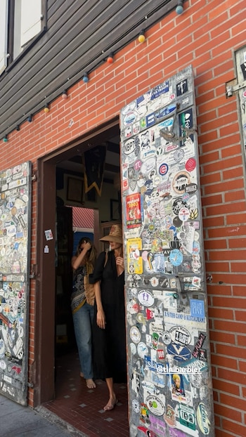 Juliana Awada y una mujer joven, ambas descalzas, entran a un edificio de ladrillos rojos con puertas de madera cubiertas de pegatinas de colores