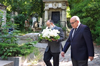 Dois homens vestidos de manto andando por um cemitério; um carregando um grande buquê de flores brancas. Ao fundo há muitas lápides e plantas