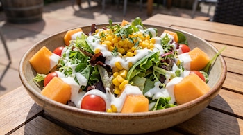 Cuenco con ensalada de hojas verdes, trozos de melón, tomates cherry, maíz dulce y salsa blanca sobre una mesa de madera.