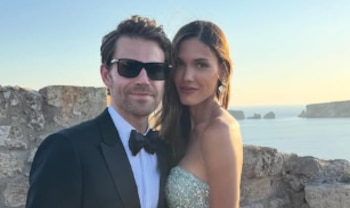 Un hombre con esmoquin y gafas de sol posa junto a una mujer con vestido brillante cerca de un muro de piedra frente al mar