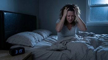Mujer con expresión angustiada sentada en la cama a oscuras, agarrándose la cabeza. Reloj digital marca 3:19 AM en mesita de noche. Hay una ventana.