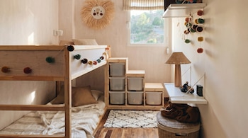 Habitación infantil con litera de madera, ropa de cama a rayas, estanterías con cajas, ventana soleada, adorno de sol en pared, lámpara de mimbre y alfombra.