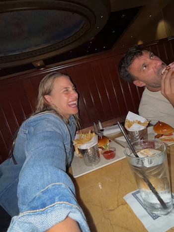 La influencer Julieta Puente riendo, con su esposo Facundo Miguelena comiendo una hamburguesa, en un restaurante con platos de comida y bebida en la mesa