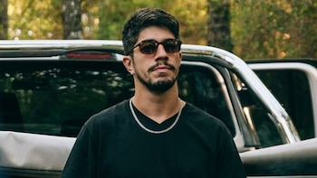 Primer plano de un hombre joven con barba, bigote y gafas de sol oscuras, vistiendo una camiseta negra y una cadena plateada. Detrás, parte de un vehículo y árboles borrosos