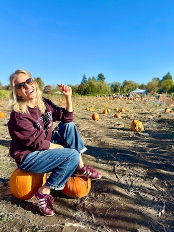 Una mujer con gafas oscuras y sudadera se sienta sobre una calabaza en un terreno de cultivo con calabazas dispersas bajo el cielo azul