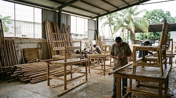 Trabajadores construyen sillas de bambú sobre mesas de madera dentro de un taller industrial con cañas apiladas junto a la pared.
