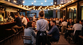 Camarera con bandeja de espaldas, clientes en mesas y barra de bar, botellas, copas, luces colgantes, edificio de fondo, noche.
