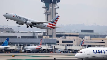 Un avión blanco de American Airlines con cola roja, blanca y azul despega en LAX, con otros aviones y terminales de aeropuerto visibles, y una torre de control al fondo