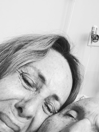 Primer plano en blanco y negro de Evelyn Von Brocke, ojos cerrados y expresión de dolor, apoyada en un hombre mayor. Parte de un equipo médico es visible