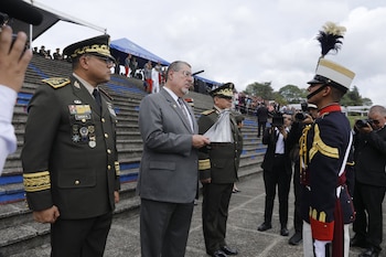 Tres hombres, dos de ellos oficiales del ejército uniformados y uno de traje, observan a un cadete con uniforme de gala en una ceremonia al aire libre