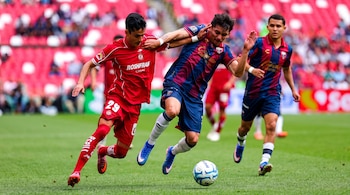 Dos jugadores de fútbol, uno con camiseta roja y otro con camiseta azul y roja, forcejean por el balón en el césped. Un tercer jugador y tribunas rojas con espectadores.