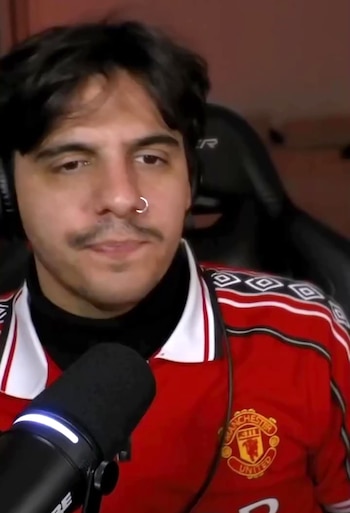Hombre con bigote, aro en la nariz y camiseta deportiva roja, situado detrás de un micrófono de estudio