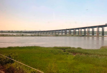 Puente largo de múltiples pilares sobre un río con aguas tranquilas y orillas cubiertas de vegetación. Cielo despejado con un avión distante en el horizonte
