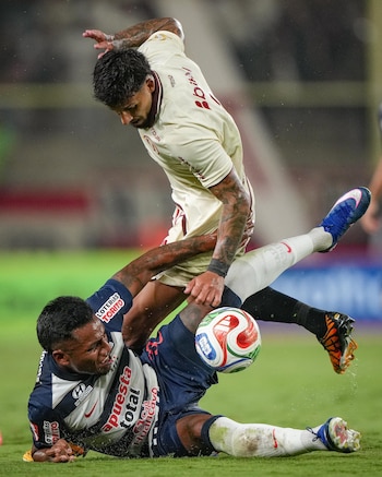 Dos futbolistas en acción intensa en un campo mojado. Un jugador con camiseta crema patea el balón mientras el otro, con camiseta a rayas, cae al suelo