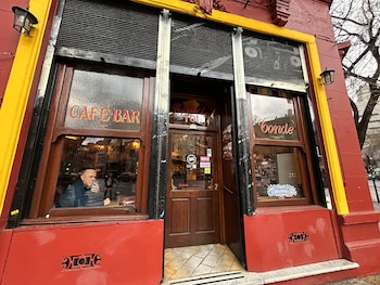 Fachada de edificio rojo con marcos amarillos, ventanas de madera, puerta de madera, letrero de Café Bar, hombre en el interior, dos faroles