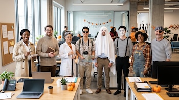 Ocho personas disfrazadas de detective, científico, fantasma y mimo, entre otros personajes, posan en una oficina con calabazas decorativas.