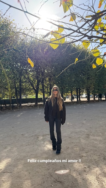 Una joven con gafas de sol y ropa oscura camina por un parque con árboles, bajo el texto escrito "Feliz cumpleaños mi amor !!"