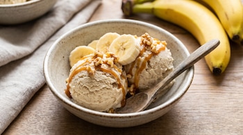 Dos bolas de helado en un tazón con rodajas de plátano, nueces, jarabe de caramelo y una cuchara de madera junto a plátanos enteros.
