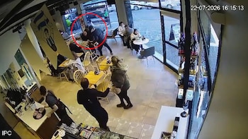 Interior de un café con mesas y sillas. Un hombre agrede a dos personas en una mesa, mientras otros clientes observan. Círculo rojo enmarca la acción
