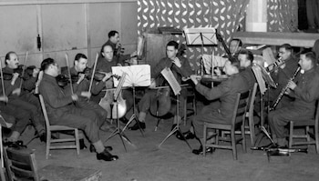 Imagen monocromática de una orquesta de hombres en uniforme tocando violines, un violonchelo, un trombón y un clarinete en una habitación