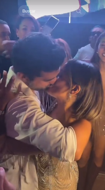 Homem e mulher se beijando intimamente, cercados por outras pessoas em festa noturna com luzes coloridas e pouca luz