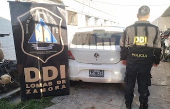 Un agente de espaldas con uniforme policial negro frente a un coche Volkswagen hatchback blanco y un cartel de la DDI Lomas de Zamora